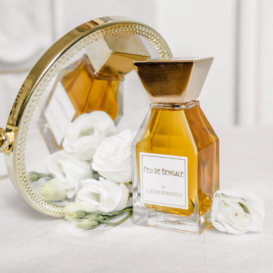 Das Flakon des exklusiven Nischenparfums Feu de Bengale von der Marke Lesquendieu auf einem goldenen Farnblatt neben einem Spiegel und weissen Rosen